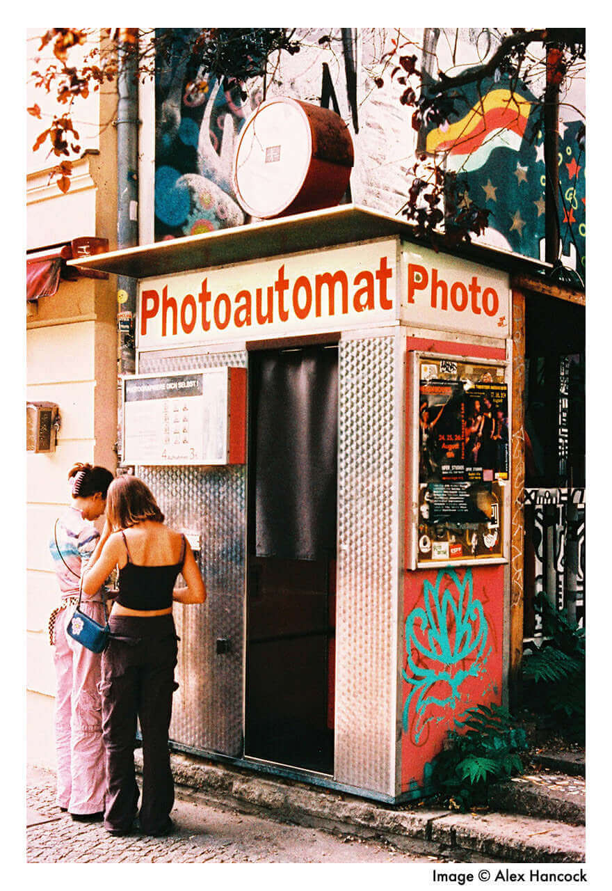 Two people using a vintage photo booth labeled 'Photoautomat' in a vibrant urban setting.