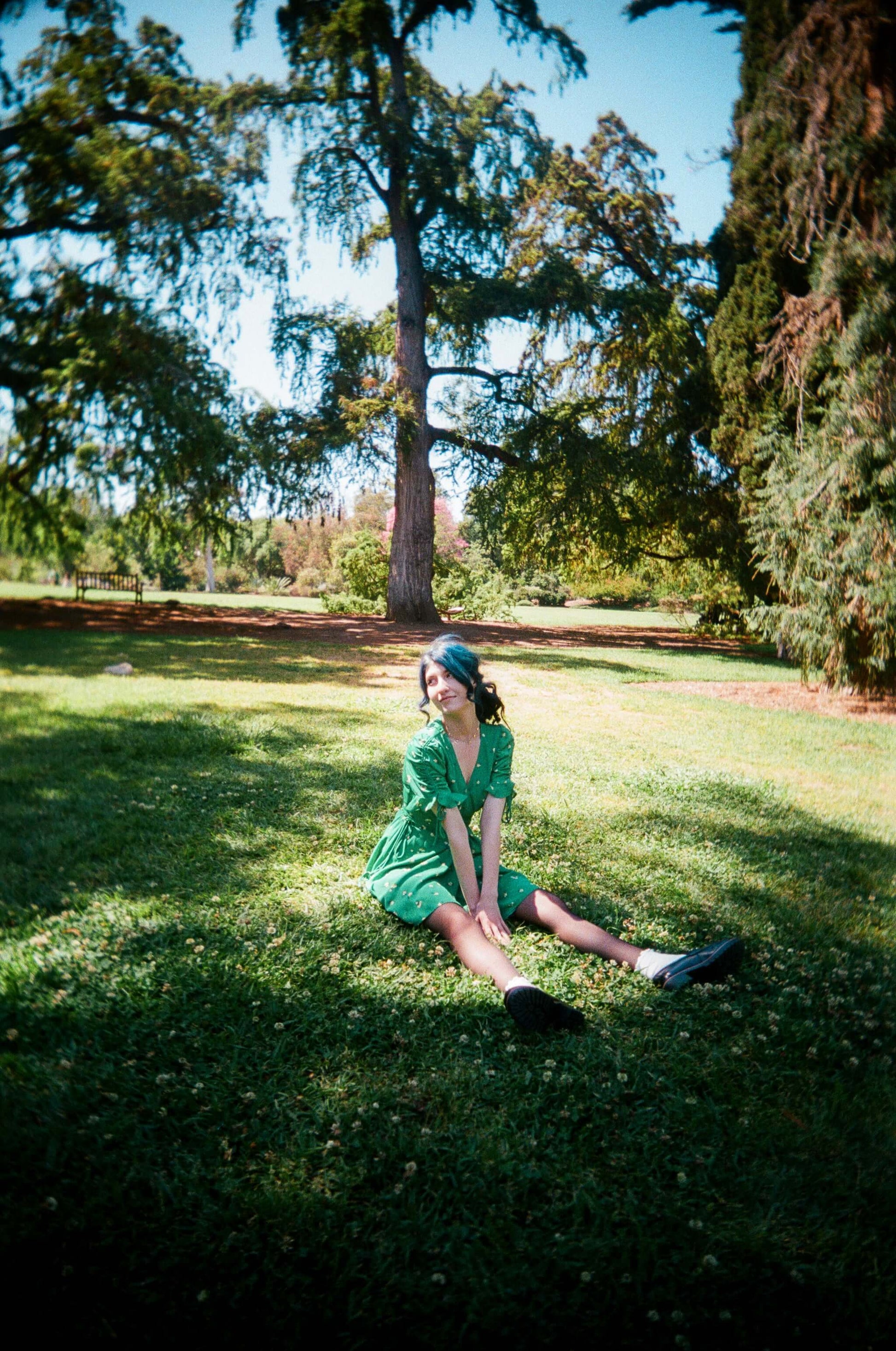 Smiling woman in a green dress sitting on grass, enjoying a sunny day outdoors.