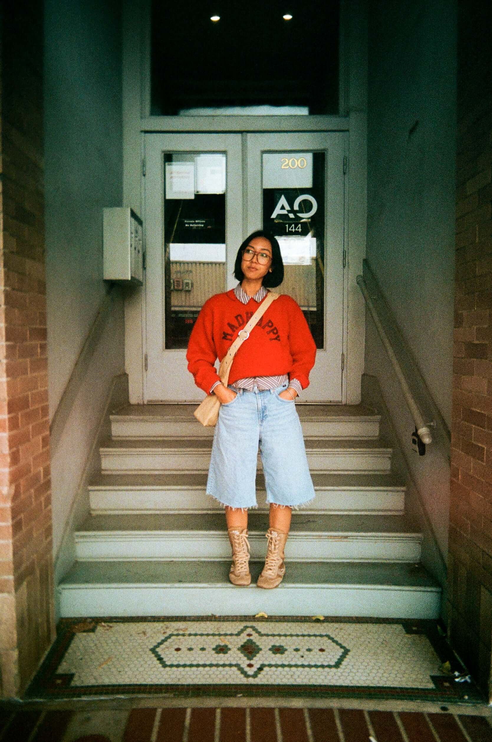 Person in red sweatshirt and denim shorts standing on stairs outside a building entrance.