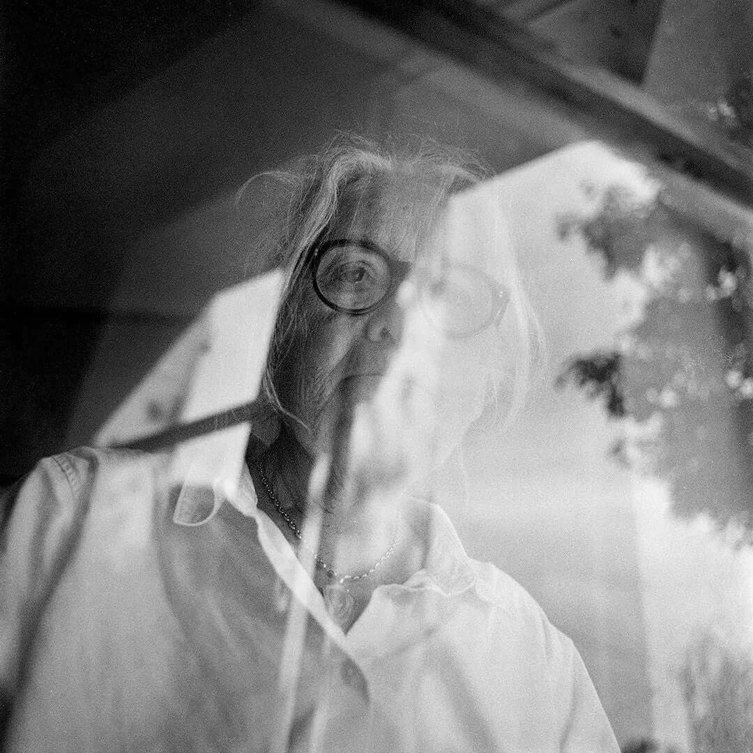 Black and white photograph captured on Bergger Pancro 400 ISO film, depicting an elderly woman seen through a glass window with reflections.