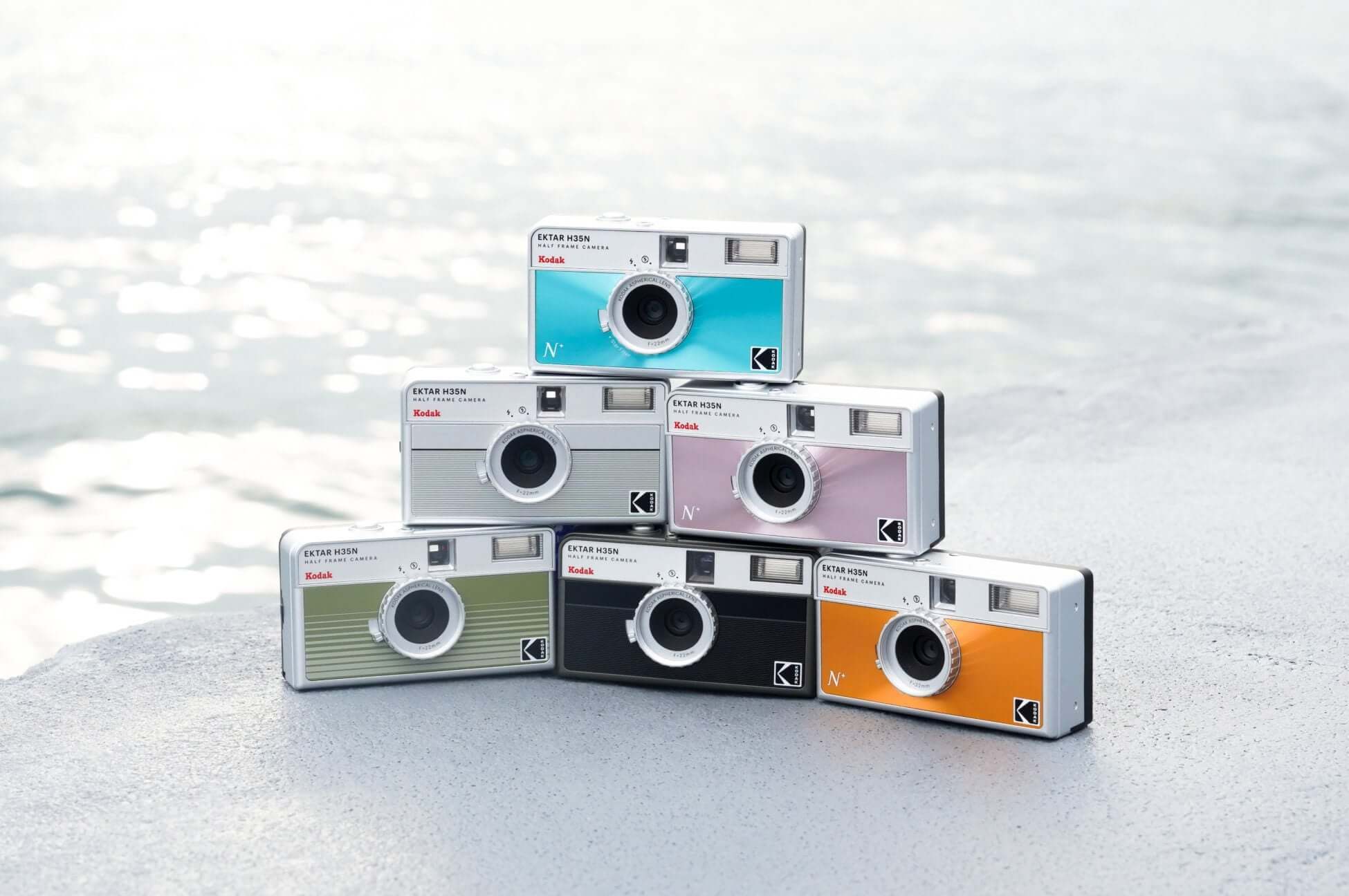 Retro-style Kodak cameras in various colors stacked on a beach, with the ocean in the background.