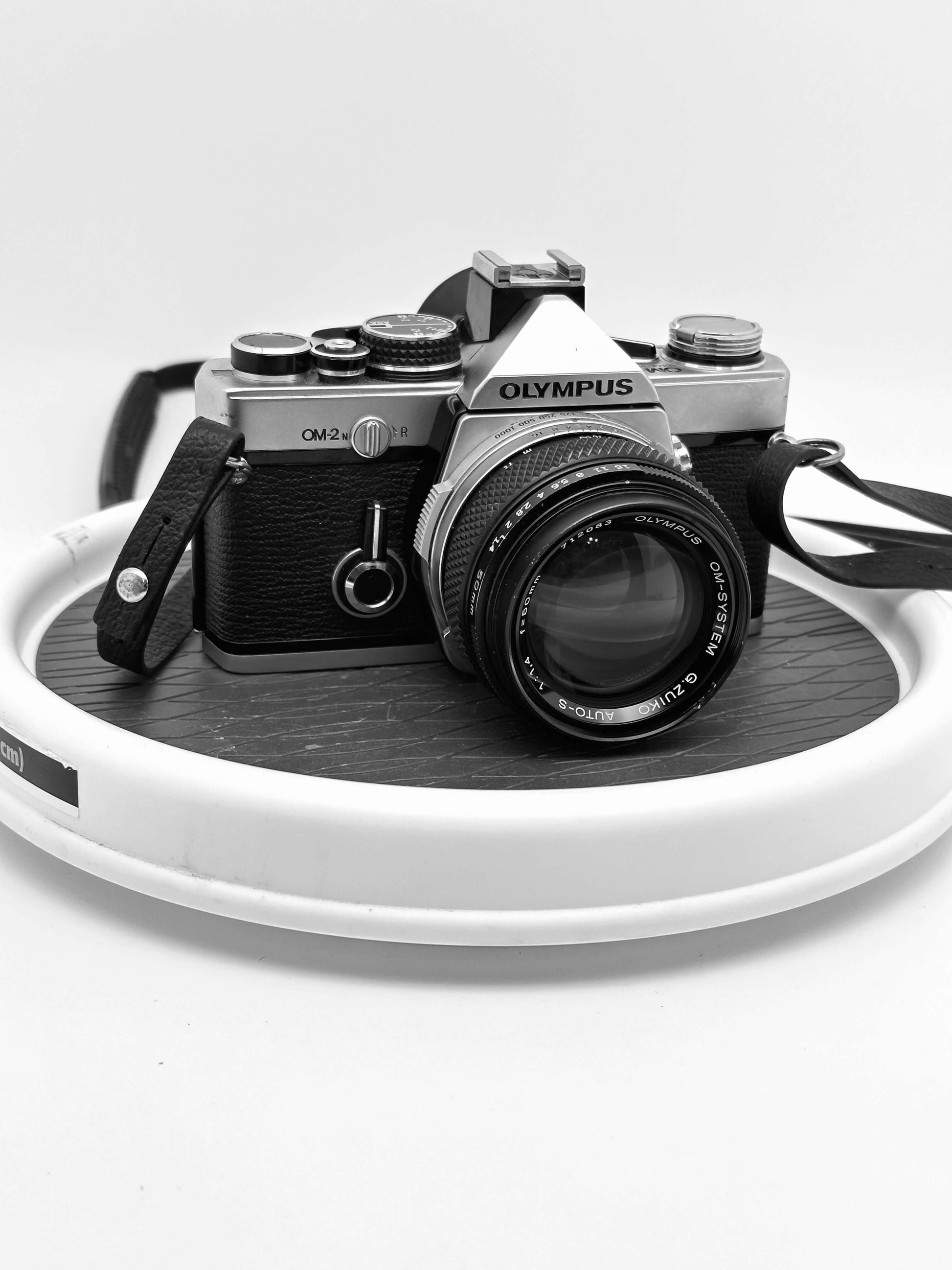 Olympus OM-2N 35mm film camera with 50mm f/1.4 Zuiko lens displayed on a white circular platform.