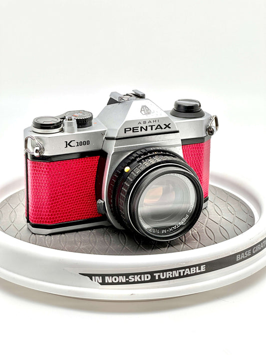 Japanese Asahi Pentax K1000 Red Used 35mm Film Camera featuring 50mm f1.7 lens on a non-skid turntable.