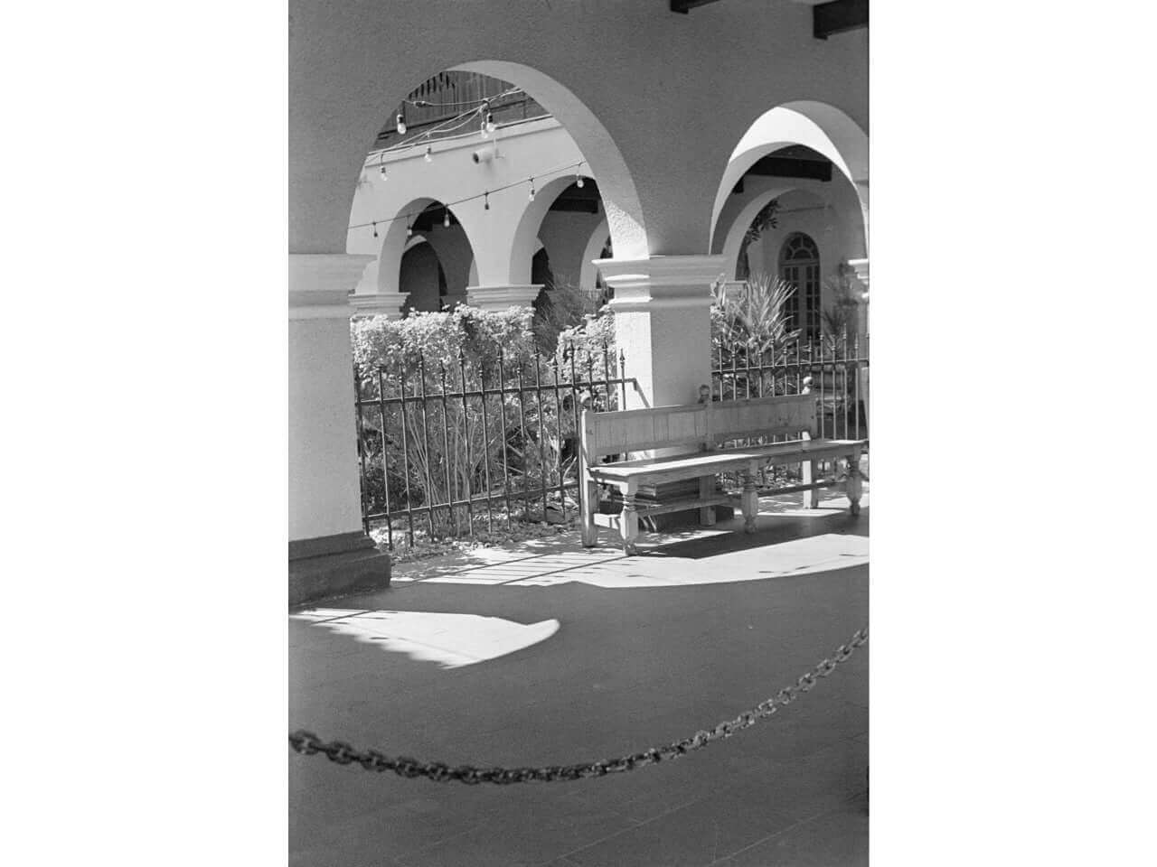 Sunlit outdoor corridor with arched walkways, potted plants, and a wooden bench.