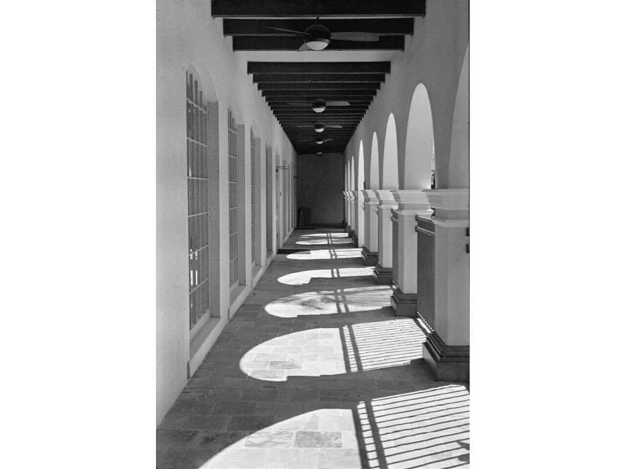 Black and white photo of a corridor featuring arches and column shadows on the tiled floor.