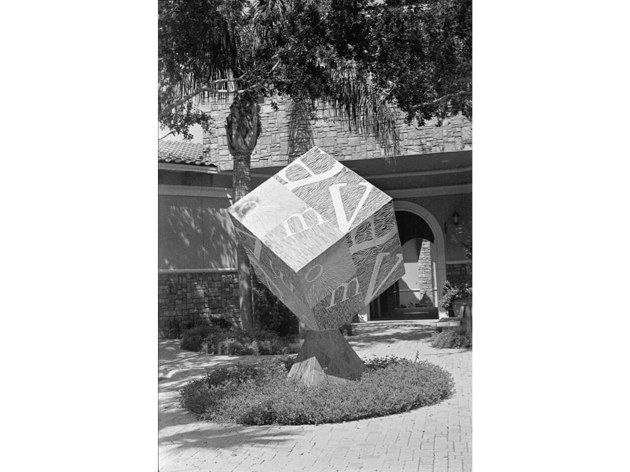 Abstract sculpture of a balanced cube with letters, displayed in a courtyard surrounded by trees and a brick building.