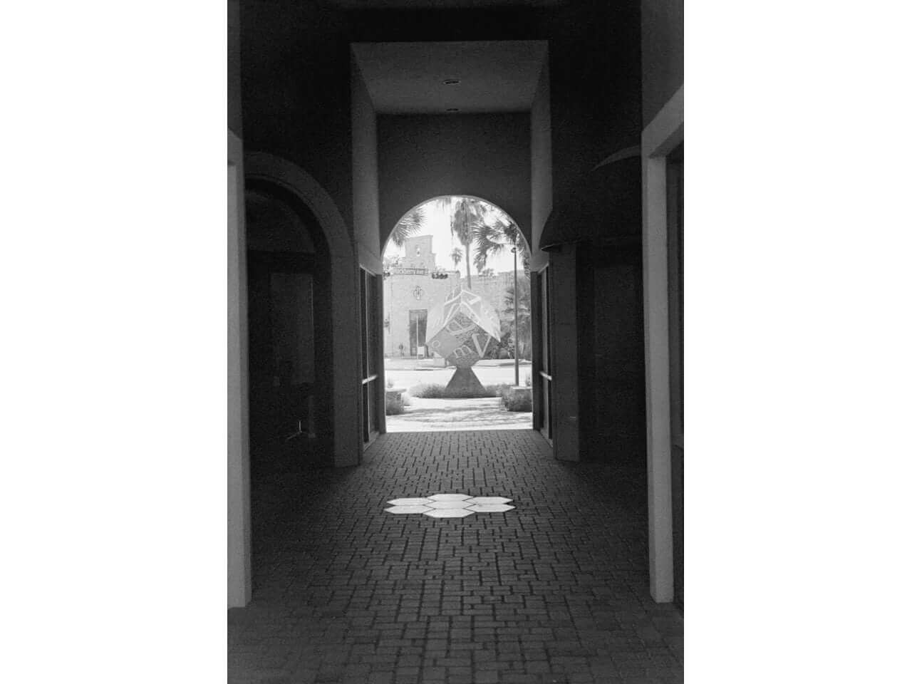 Black and white pathway with arch leading to courtyard and geometric sculpture in the distance.