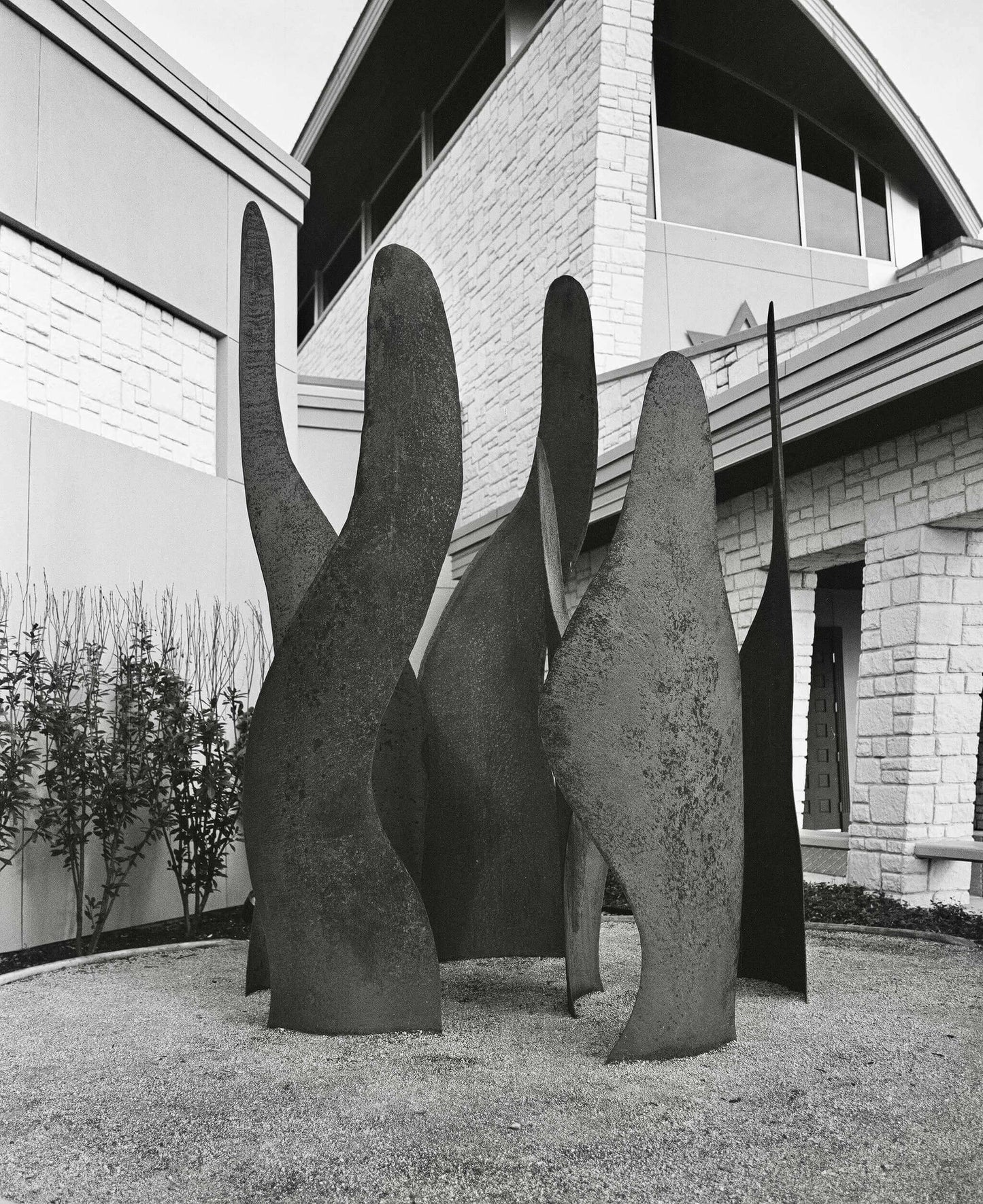 Black and white photograph of abstract metal sculptures in a landscaped area near a modern building.
