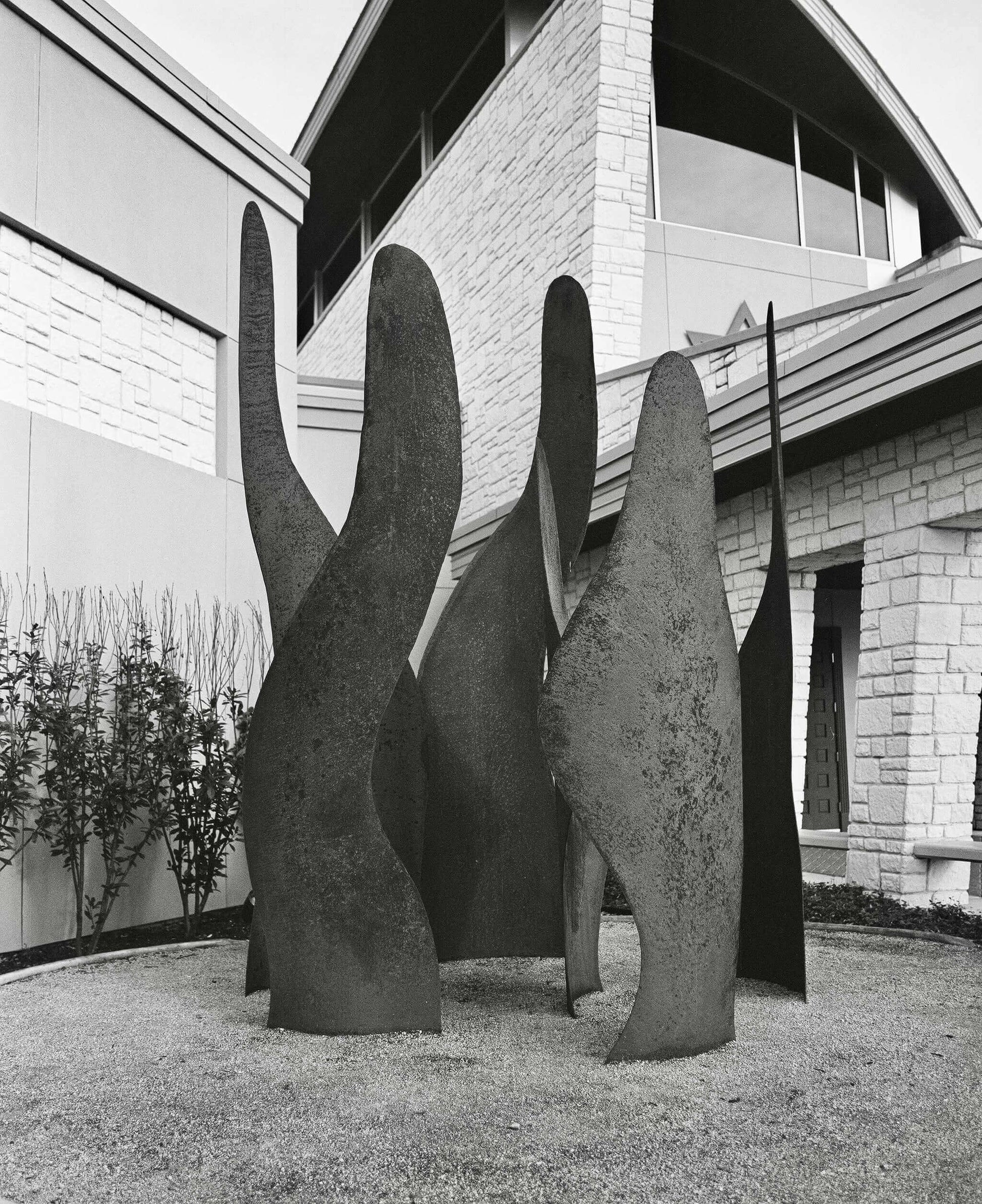 Black and white photograph of abstract metal sculptures in a landscaped area near a modern building.