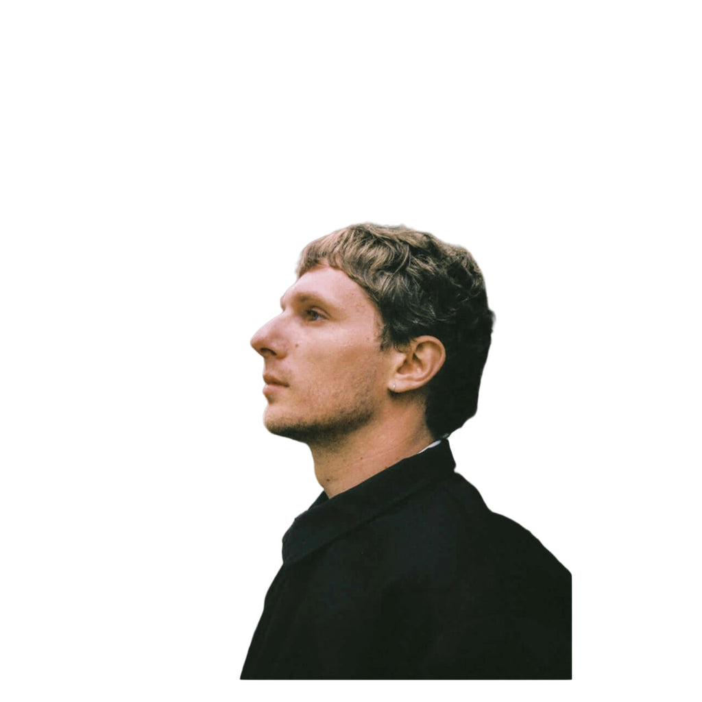 A man with short brown hair and a light stubble, wearing a black coat.