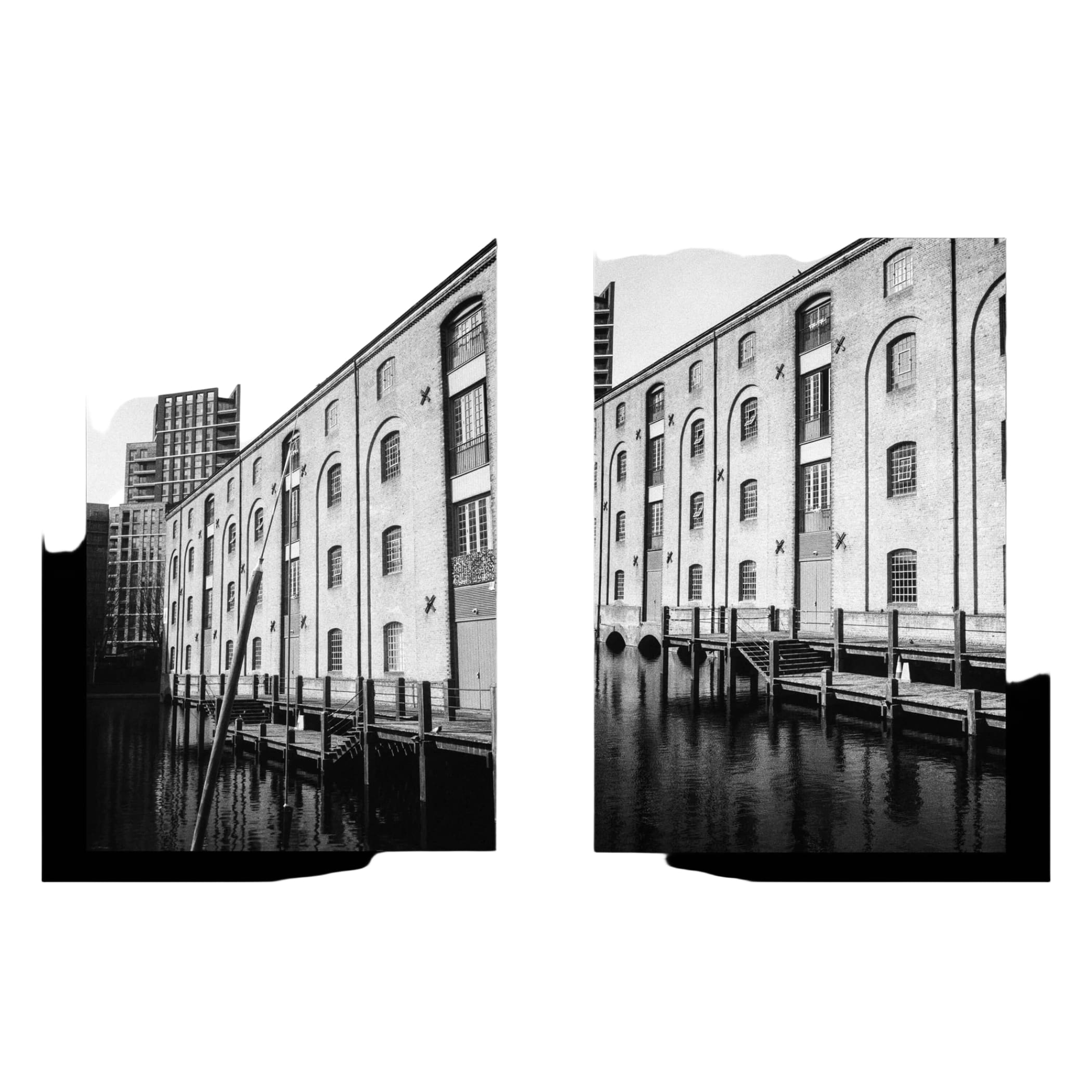 Black-and-white photograph of a historic brick warehouse building with arched windows and wooden docks along the water.