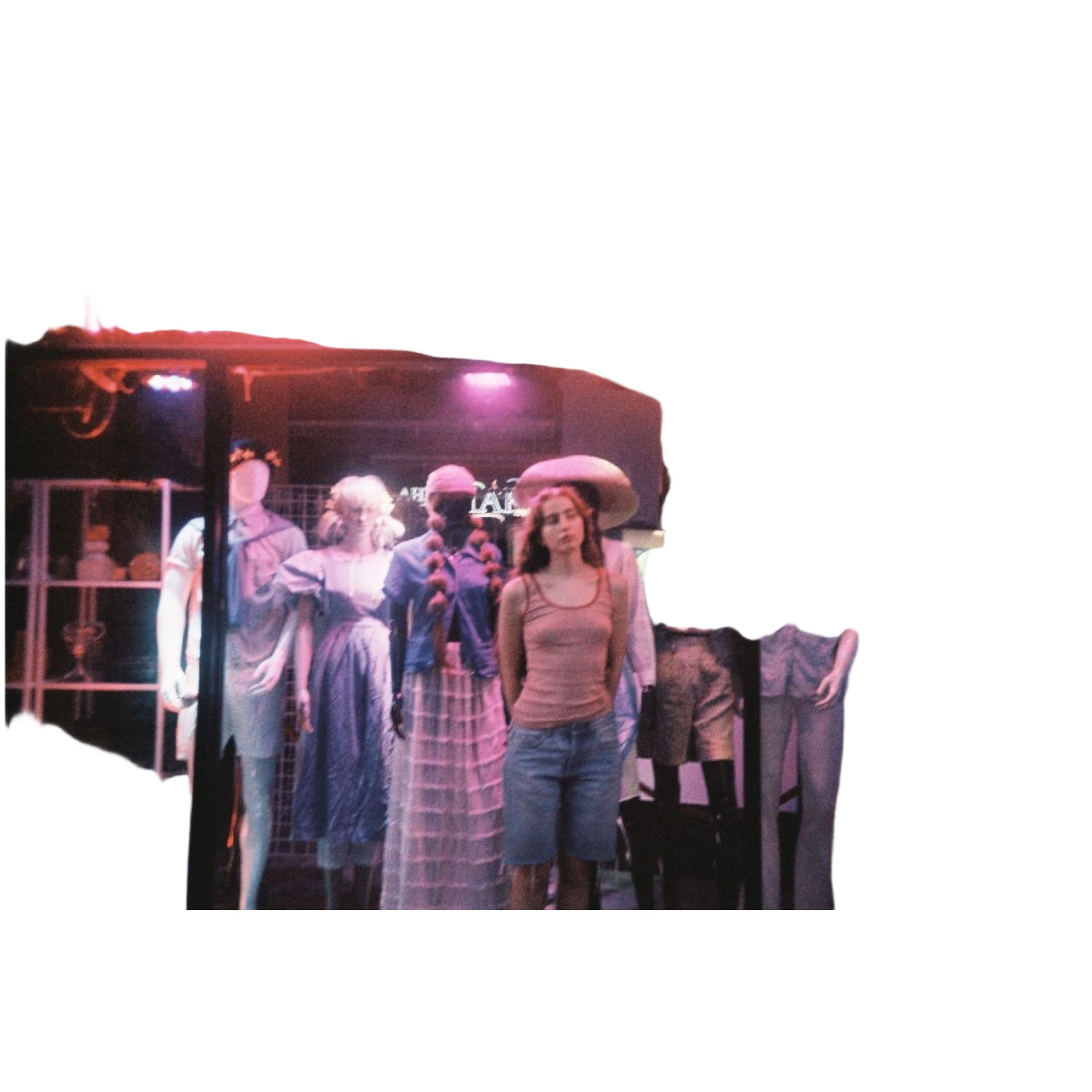 A woman stands in front of a thrift store window display, wearing a tan tank top and blue denim shorts.