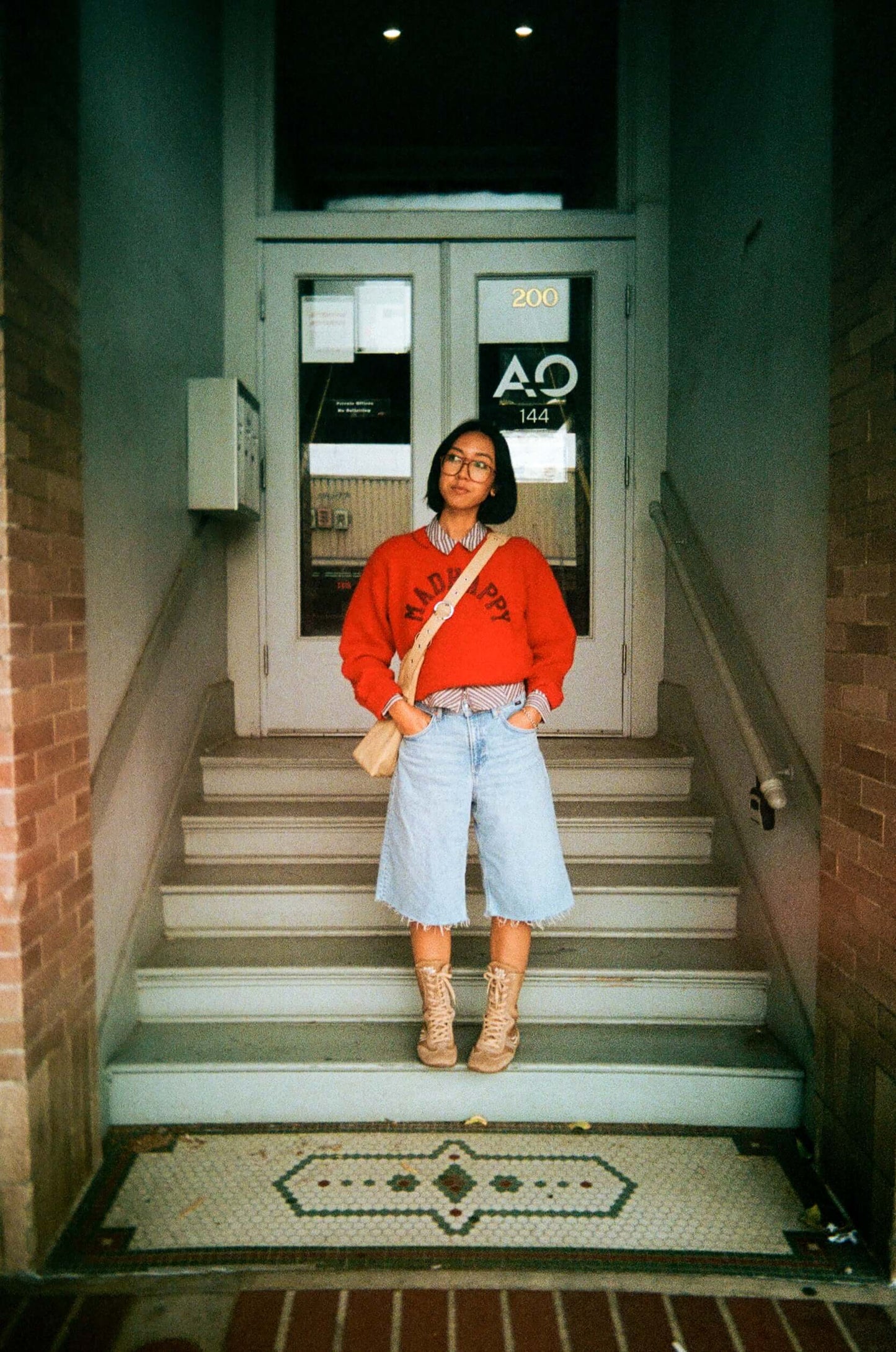 Person in red sweatshirt and denim shorts standing on stairs outside a building entrance.