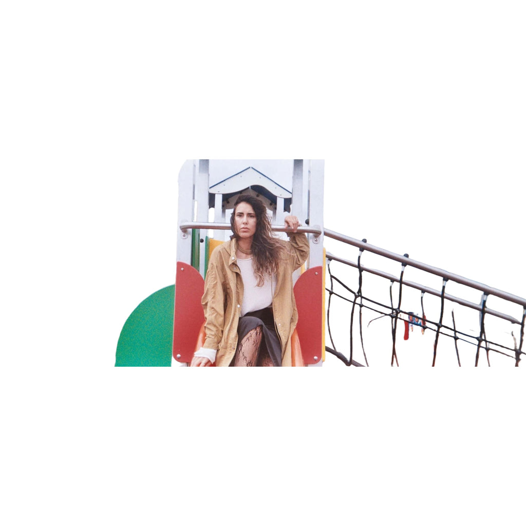 A woman with long, wavy brown hair stands on a colorful playground structure wearing an oversized tan jacket over a white top and patterned leggings.