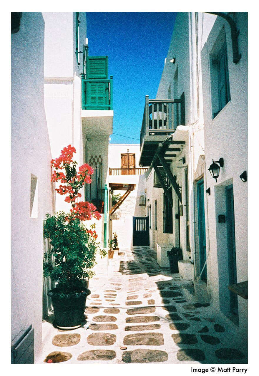 Charming narrow street with white buildings and vibrant flowers in a Mediterranean village under a bright blue sky.