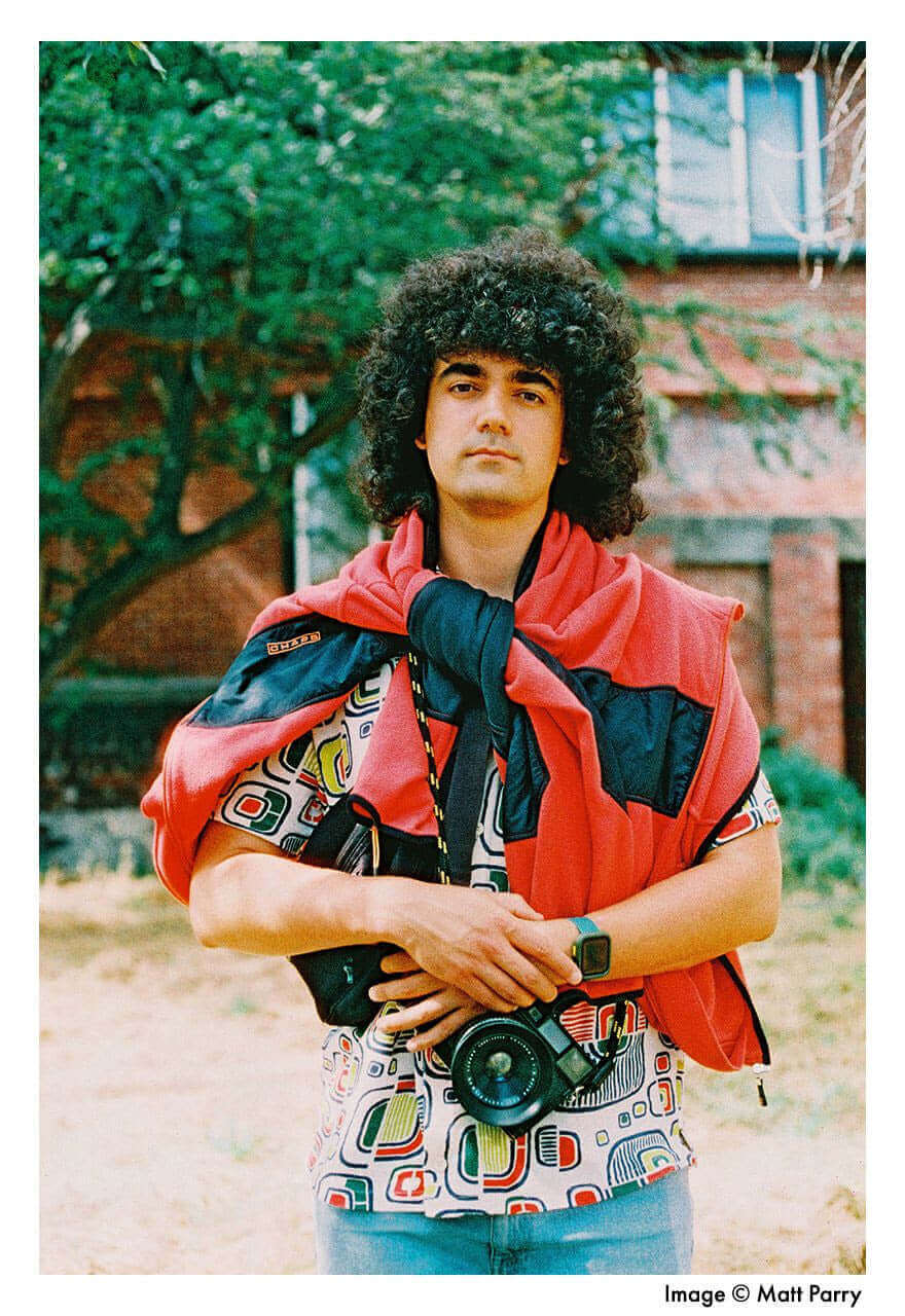 Person with curly hair and colorful clothing holding a camera outdoors in front of a brick building.