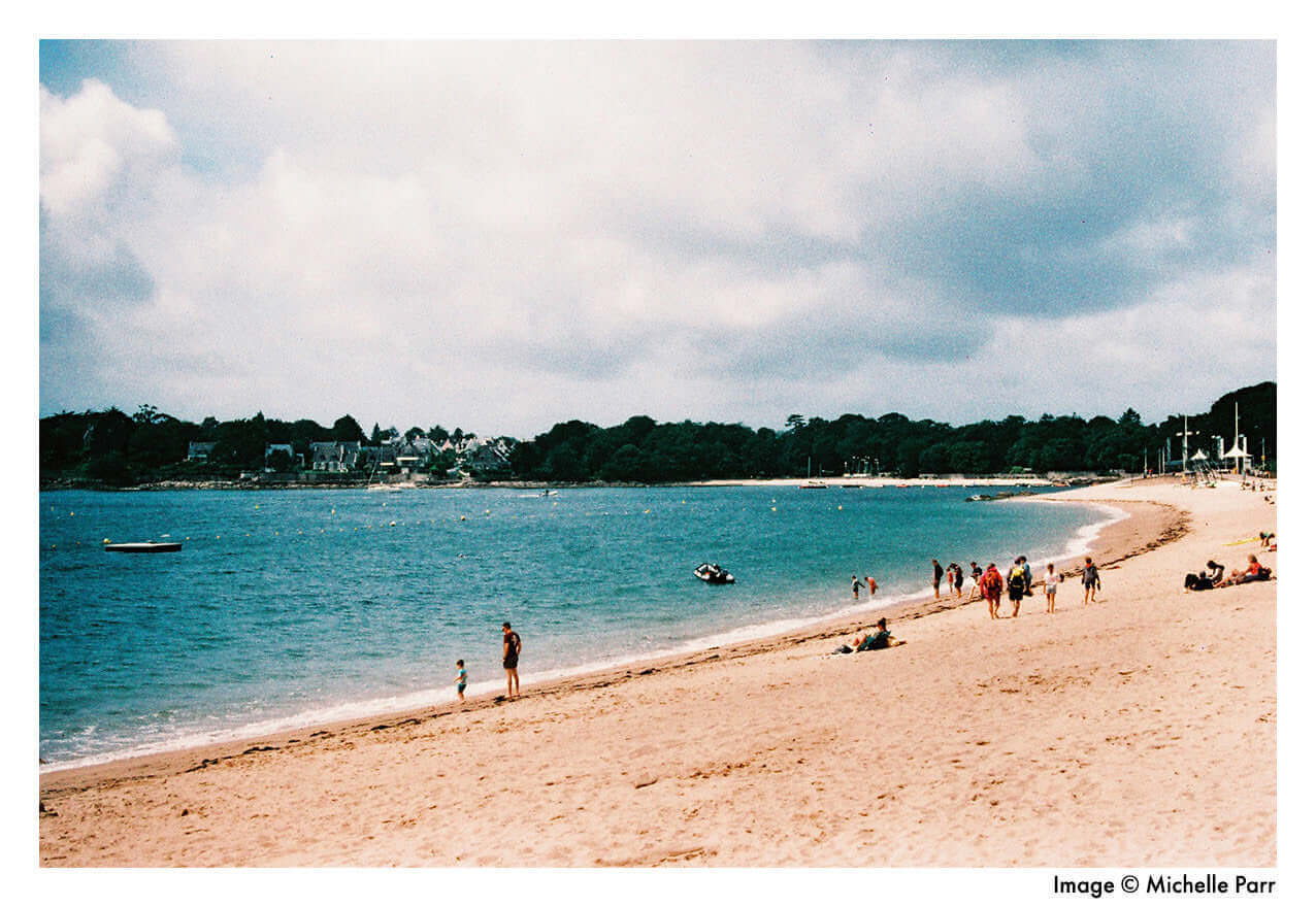 Scenic beach with people enjoying a sunny day, blue waters, and picturesque coastal town in the distance.