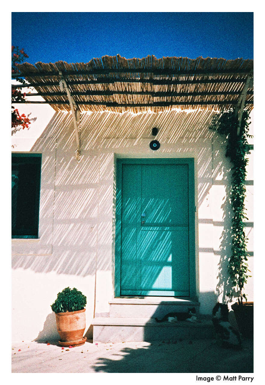 Sunlit turquoise door with canopy casting shadows on white exterior wall and potted plant underneath.