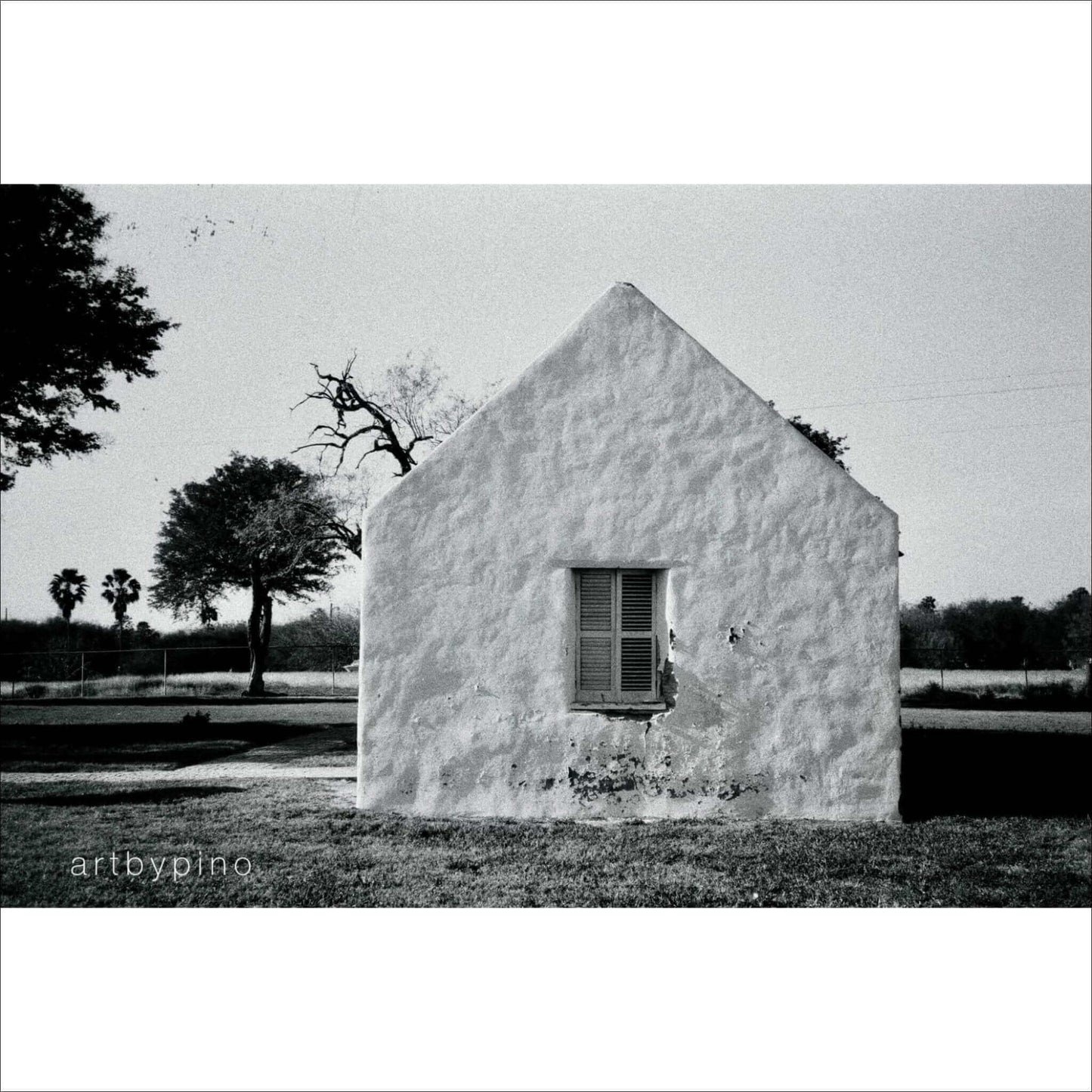 Black and white photo of a simple white house with a window, surrounded by trees and open land.