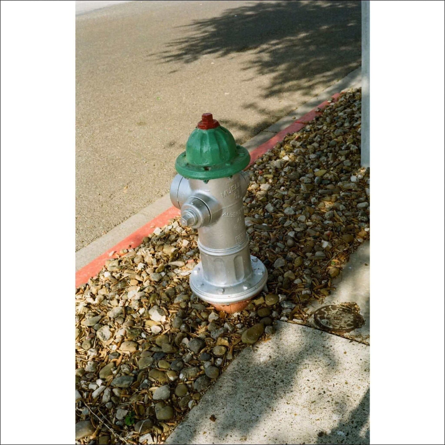 Silver fire hydrant with green and red top placed on a gravel area next to a street with a shadow of a tree in the background.