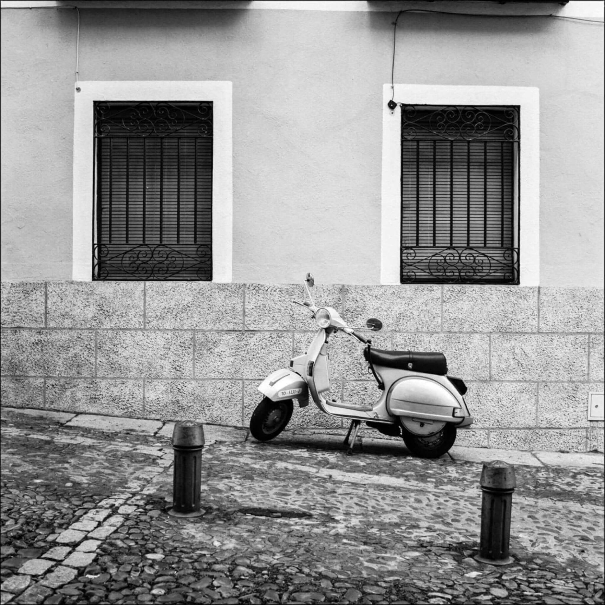 Classic Vespa scooter parked on a cobblestone street between two shuttered windows in black and white photo.