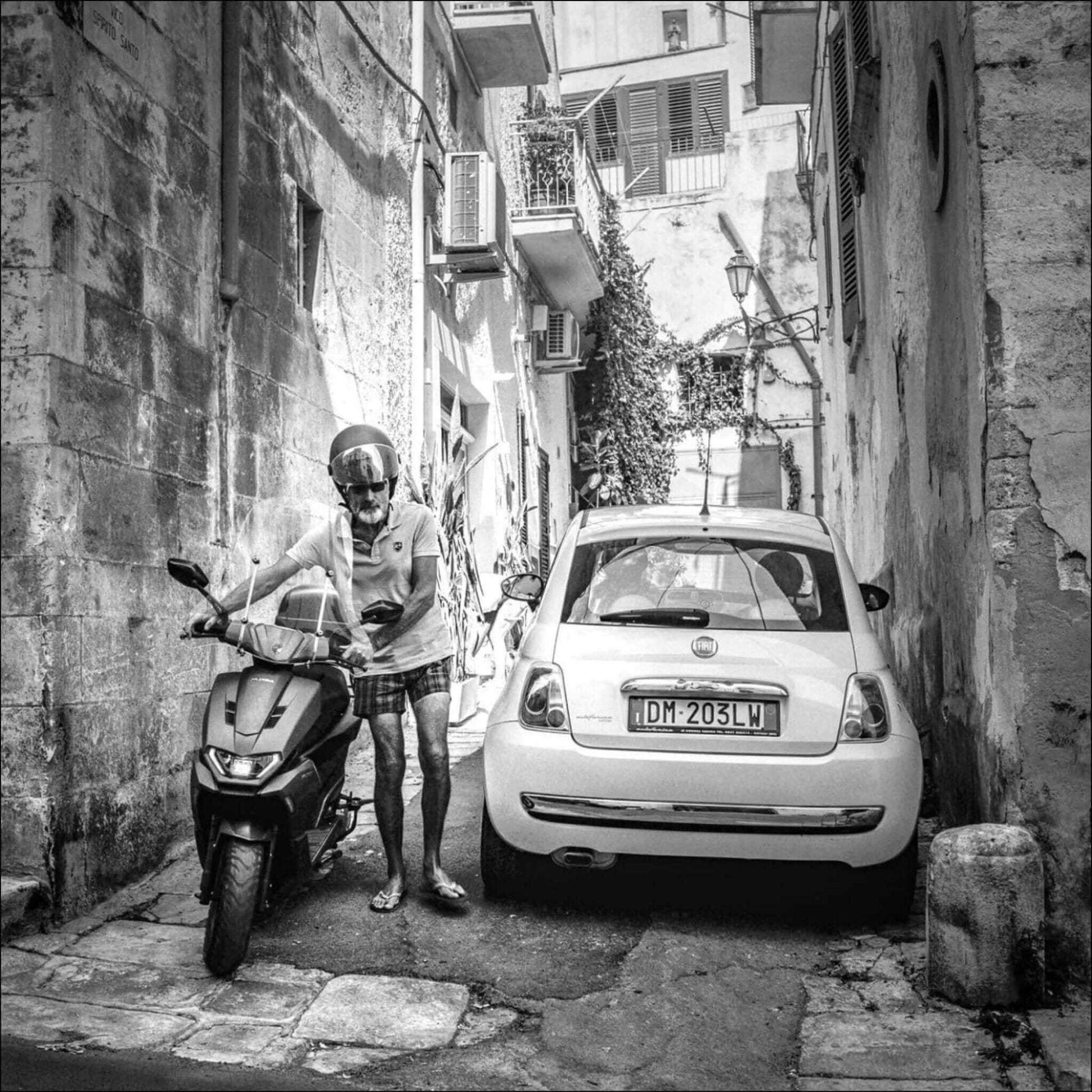 Black and white photo of a man with a helmet checking his scooter beside a parked white car in a narrow alley.