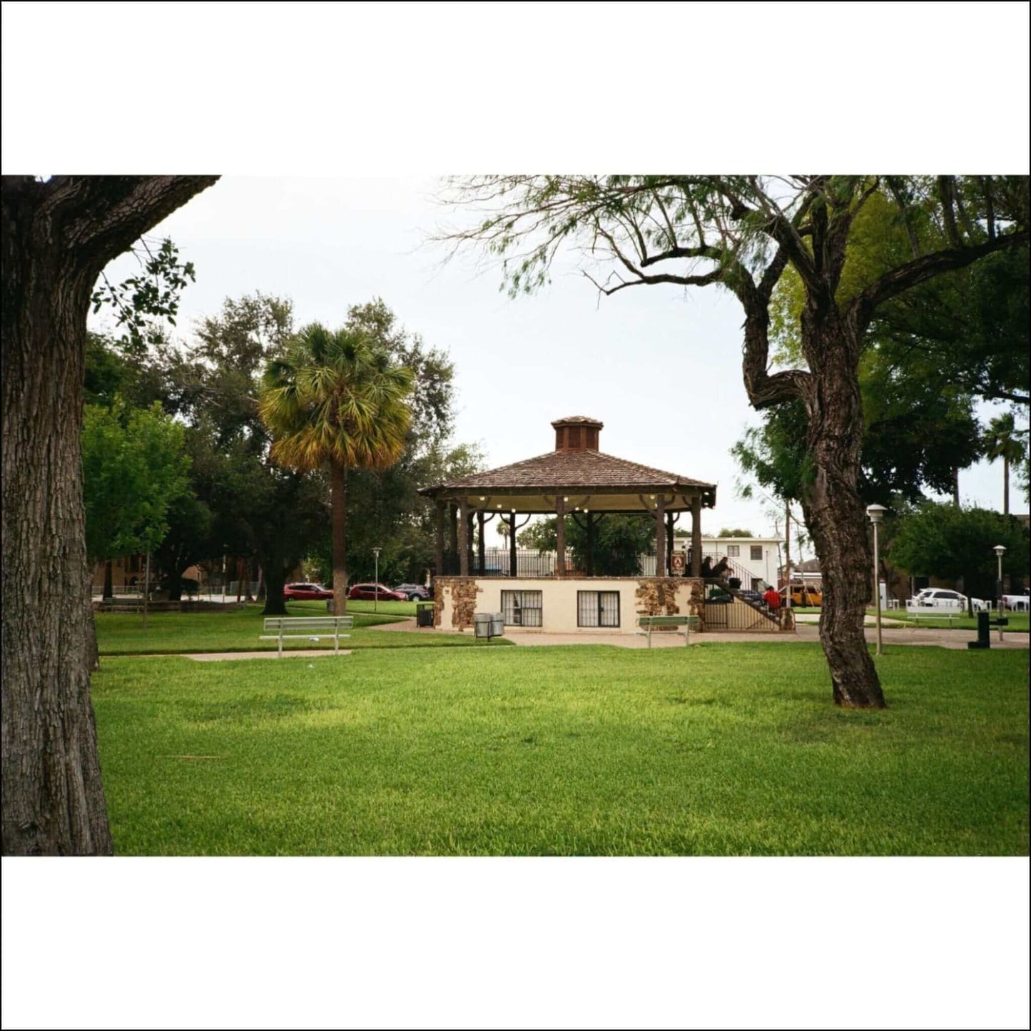 Gazebo in a scenic park with lush green grass, trees, and palm trees in the background on a calm day