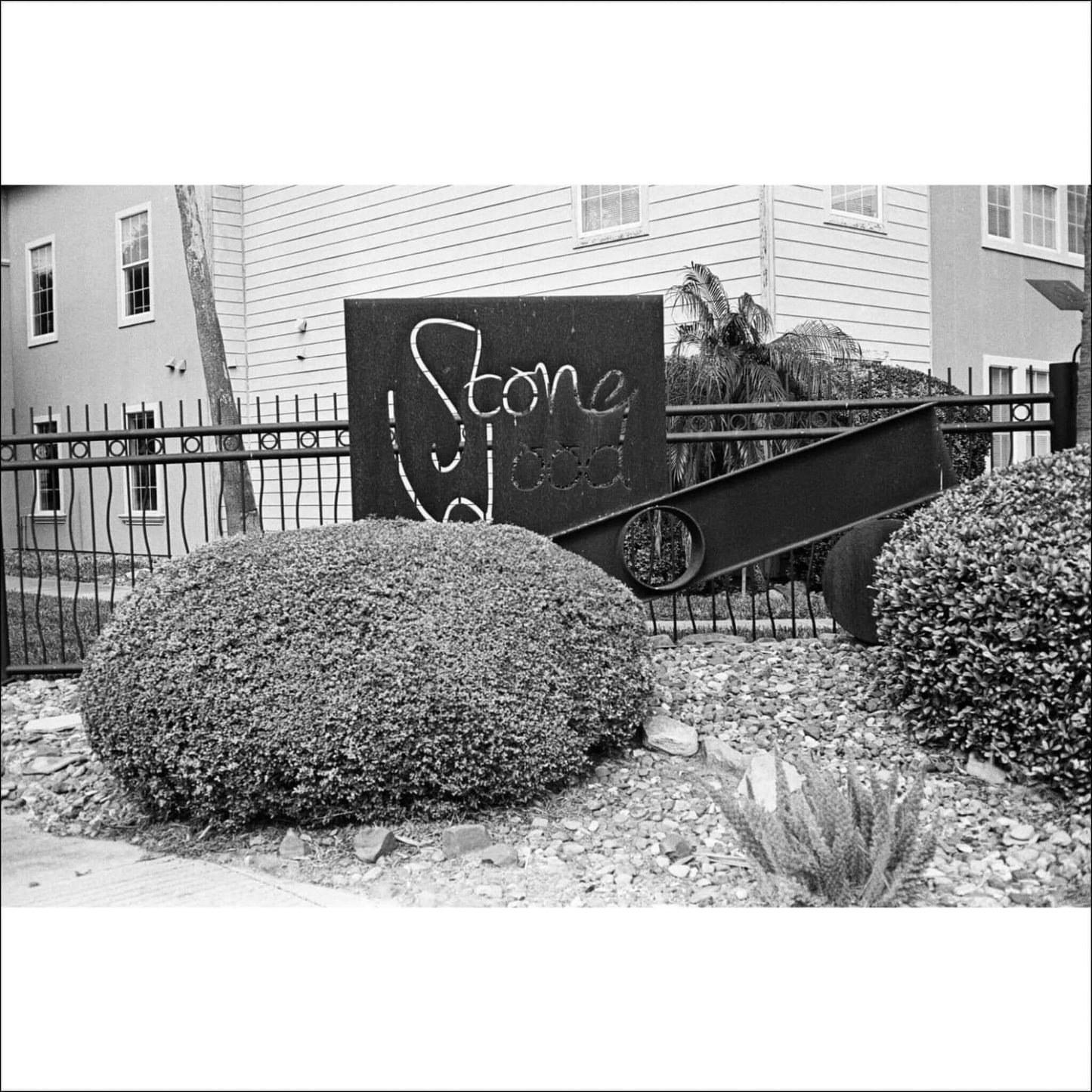 Black and white photo of a landscaped area with bushes and a "Stone Wood" sign in front of a fence and a building.