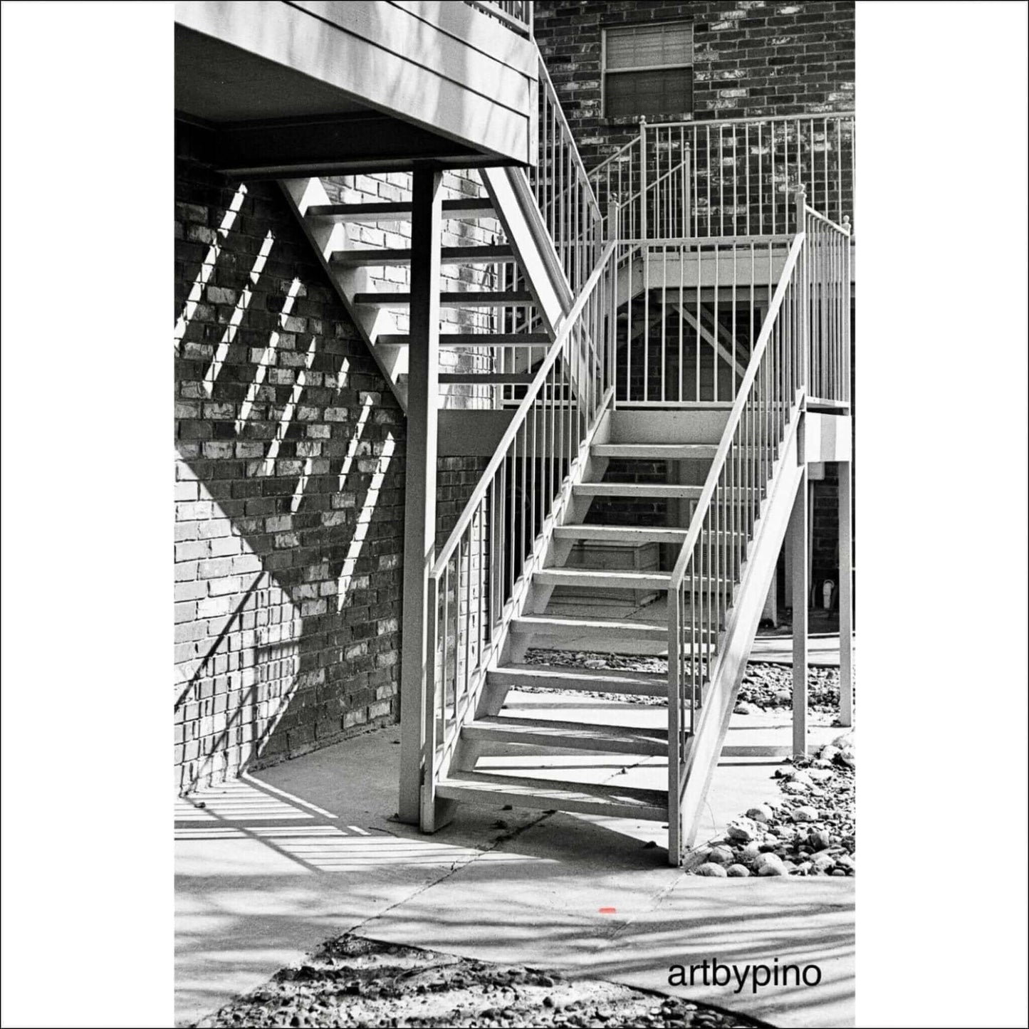 Black and white photo of outdoor staircase with metal railings and brick walls, shadows creating geometric patterns. Art by Pino.