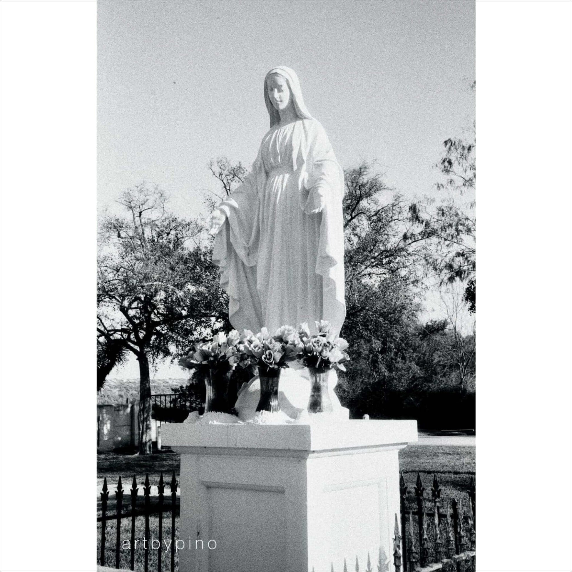 Statue of a woman in white robes surrounded by flowers in an outdoor setting with trees in the background