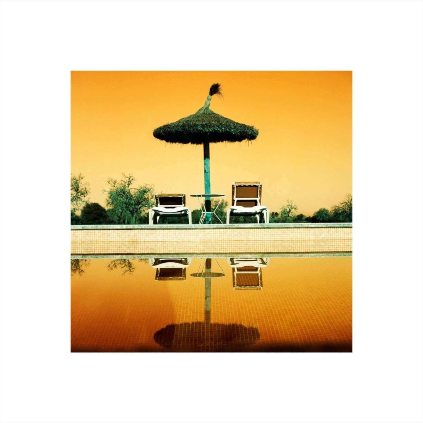 Poolside scene with two lounge chairs and a thatched umbrella reflecting in tranquil water at sunset.