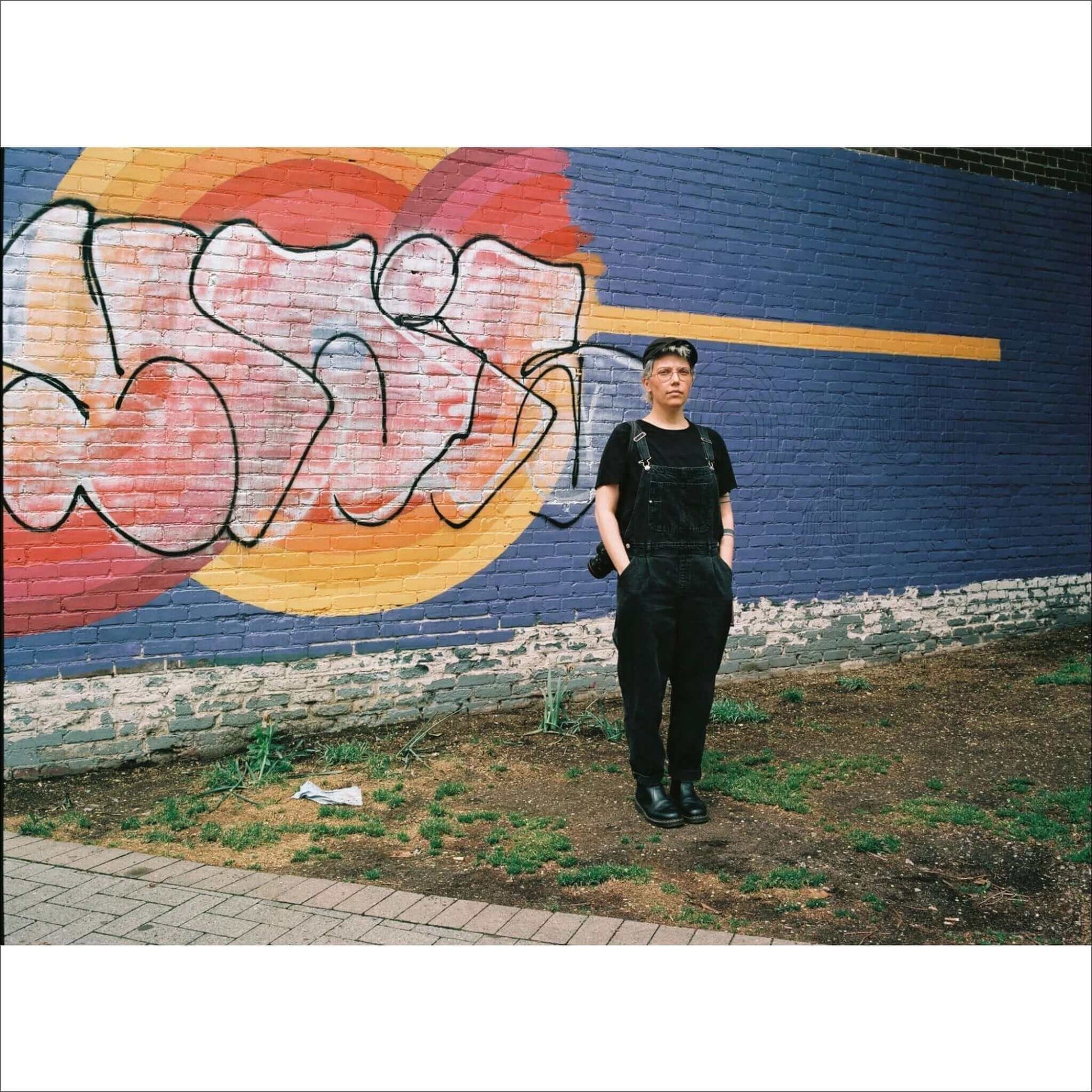 Person standing in front of a colorful graffiti wall while holding a camera, dressed in black overalls.